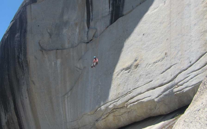 a person wearing safety gear is suspended by ropes mid air against the backdrop of a large smooth rock wall